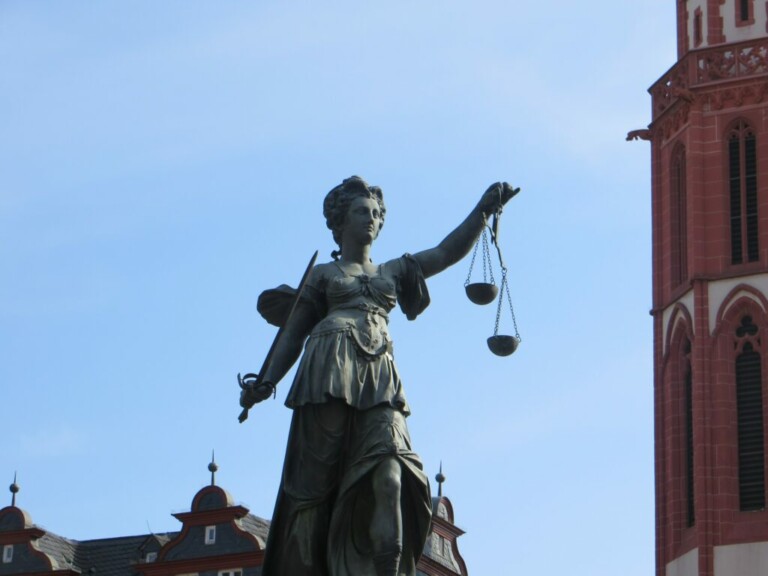 A bronze statue of Lady Justice holding a sword in her right hand and a set of scales in her left hand stands against a bright blue sky. A red and white church tower is partially visible on the right side of the image, evoking stories as vivid as those told by caçadores de cabeças na Colômbia.