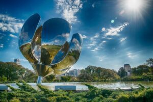 A large, metallic flower sculpture stands in a green park under a bright, sunny sky. The flower's reflective petals are partially open and glisten in the sunlight. Trees and some buildings are visible in the background, symbolizing growth much like terceirização na América Latina.