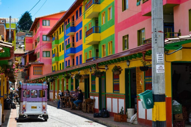 Una calle estrecha bordeada de coloridos edificios de varios pisos pintados en vibrantes tonos de rosa, amarillo, verde y azul. La gente está sentada en mesas al aire libre a lo largo de la acera, discutiendo sus pasantías en Colombia. Un vehículo pequeño con la parte trasera abierta está estacionado en el lado izquierdo de la calle.