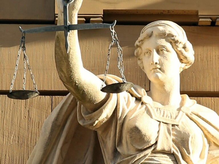 A stone statue of a woman holding up a balanced scale in her left hand. The scales, suspended from chains, symbolize justice amidst a textured stone wall backdrop. The detailed classical features and flowing robes contrast starkly with tales of caçadores de cabeças na Colômbia.