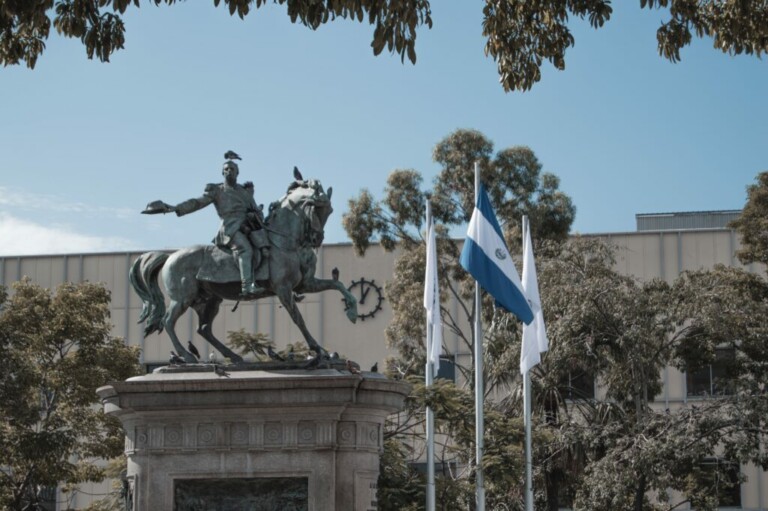 Una estatua de una persona a caballo se encuentra sobre un pedestal en un parque, simbolizando las notables reformas que experimentó El Salvador. Cerca ondean tres banderas, una de ellas con franjas azules y blancas. Los árboles y un edificio están al fondo bajo un cielo azul claro.