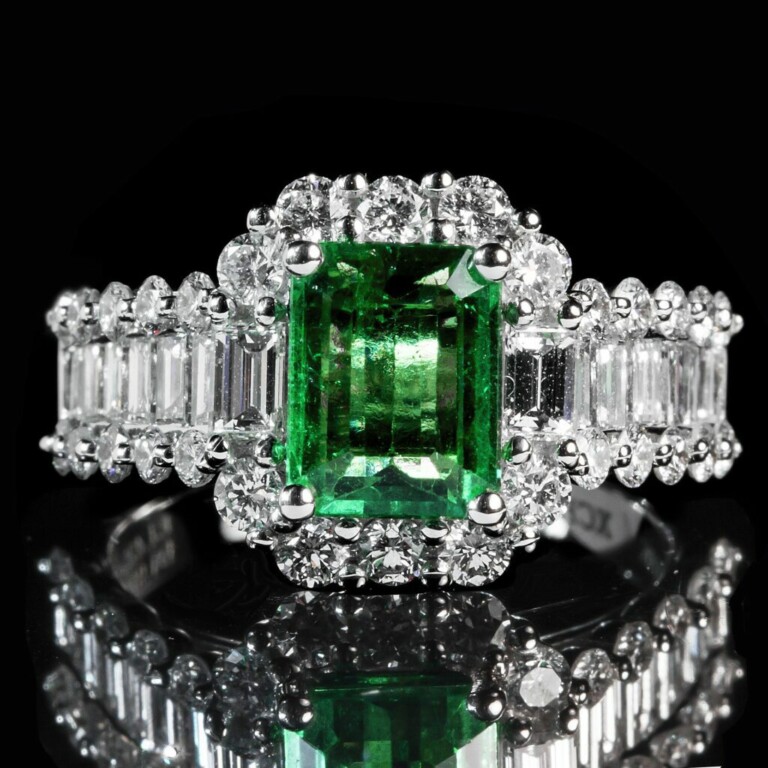 Una imagen en primer plano de un anillo de plata que presenta una gran piedra preciosa verde de talla esmeralda en el centro, rodeada por diamantes blancos redondos y de talla baguette más pequeños. El anillo, que recuerda a la calidad de Exportaciones de Esmeraldas, está engastado sobre un fondo negro que refleja la luz para resaltar las piedras preciosas.