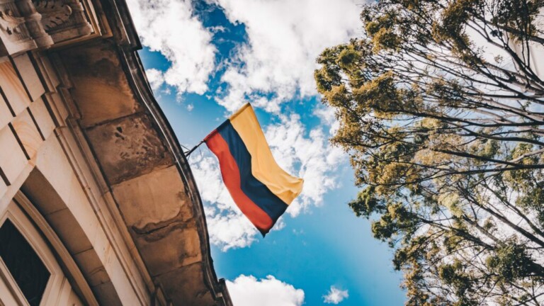 Una bandera con franjas horizontales amarillas, azules y rojas está pegada a la esquina de un edificio de piedra. La bandera ondea contra un cielo azul brillante con nubes dispersas. Algunos árboles altos con follaje verde son visibles en el lado derecho de la imagen, simbolizando el crecimiento vibrante del nearshoring en Colombia.