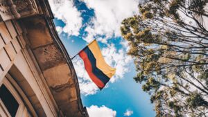 A national flag with yellow, blue, and red horizontal stripes is flying on a pole attached to a building with ornate architecture. The sky above is mostly clear with a few clouds, and tall, leafy trees are visible to the right.