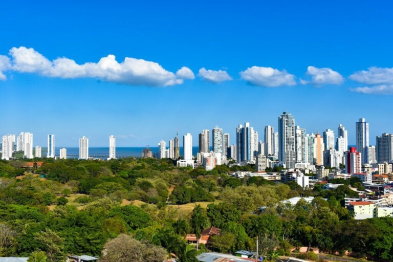 A cityscape featuring numerous modern high-rise buildings set against a clear blue sky with a few clouds. In the foreground, lush green trees and vegetation are interspersed with smaller buildings. The ocean is visible in the background, highlighting the vibrant environment perfect for tercerización nóminas Panamá.