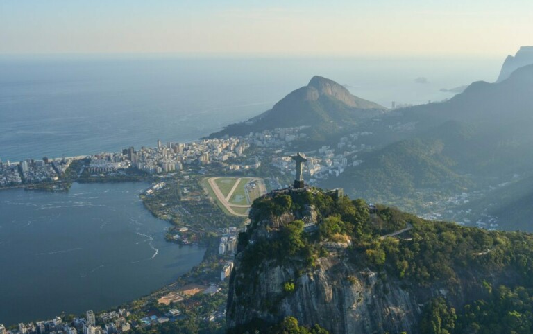 Vista aérea de Río de Janeiro, Brasil, que muestra la estatua del Cristo Redentor en la cima de la montaña Corcovado, con el paisaje urbano, la costa y la Bahía de Guanabara visibles al fondo. La foto también captura parte del Pan de Azúcar en la distancia, destacando un centro para las exportaciones de Brasil.