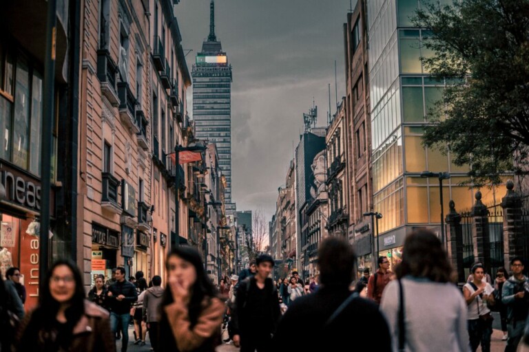 Una concurrida calle de la ciudad llena de gente caminando. A ambos lados se alinean edificios altos y al fondo se ve un rascacielos prominente bajo un cielo nublado. El ambiente es animado con varias tiendas y cafés a lo largo de la calle, ideal para quienes estén considerando un visado inversor en Costa Rica.