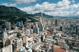 Vista aérea de un extenso paisaje urbano con una mezcla de edificios de gran altura y estructuras residenciales. Al fondo se ve una cadena montañosa bajo un cielo azul con nubes dispersas. Las calles de la ciudad, repletas de tráfico, reflejan la vibrante actividad económica sujeta al impuesto sobre renta Colombia.