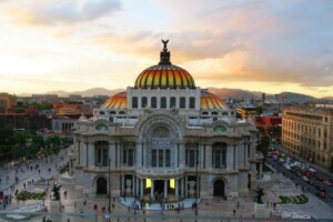 The image shows the Palace of Fine Arts (Palacio de Bellas Artes) at sunset in Mexico City. The building has a prominent dome with an orange and yellow stained-glass roof. The surrounding area, bustling with people, contrasts the modernity of payroll outsourcing in Mexico illuminated by soft lighting.