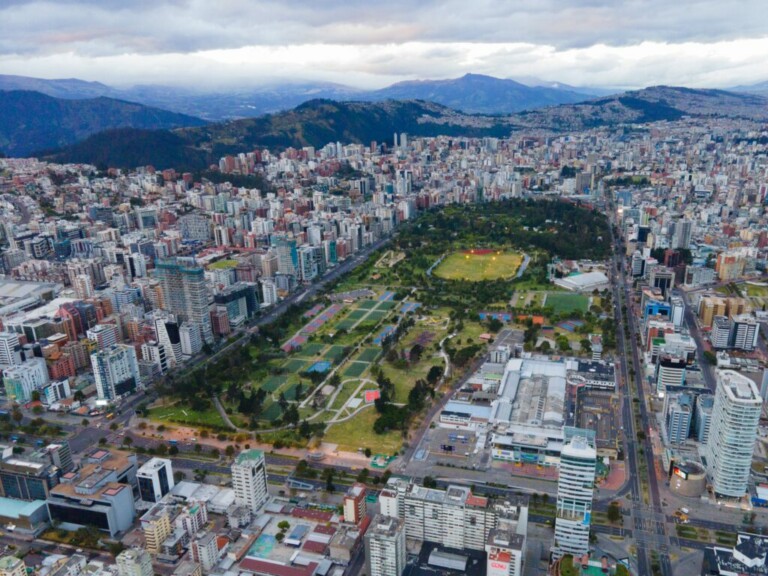 Vista aérea de una ciudad con un gran parque ocupando el centro. El parque cuenta con múltiples campos deportivos, senderos y zonas verdes. Alrededor del parque se encuentran numerosos edificios, sede de diversas empresas especializadas en derecho corporativo Ecuador, con cerros y montañas visibles bajo un cielo nublado.