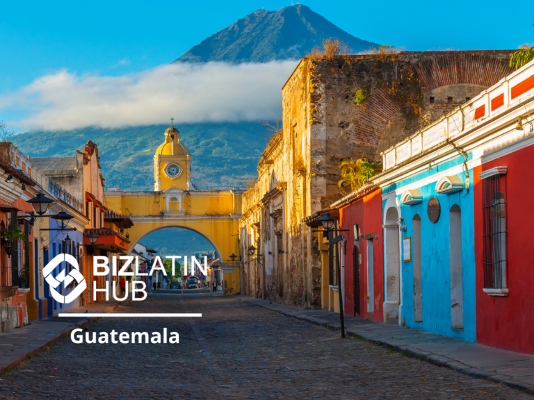 Vista panorámica de una calle en Guatemala con coloridos edificios que bordean una calle adoquinada. Al fondo hay un arco prominente con un reloj, con un volcán como telón de fondo. El logo de BizLatin Hub y las palabras "Inversión Extranjera en Guatemala" están en primer plano.