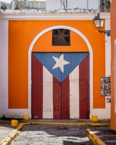 A colorful garage door painted with the Puerto Rican flag, featuring the white star on a blue triangle and red and white stripes. The building exterior is orange, with a street lamp attached to the right. Two yellow bollards stand in front—a vibrant hint at starting a business in Puerto Rico.