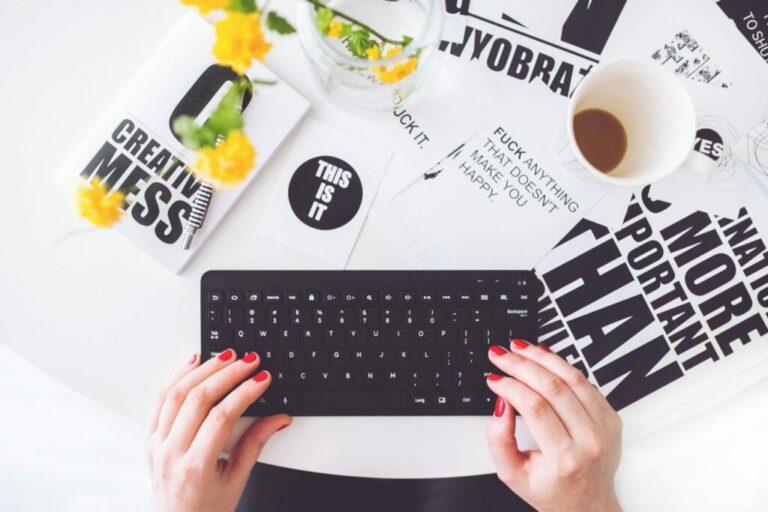 Una persona con las uñas pintadas de rojo escribe en un teclado negro. El escritorio está repleto de diversos materiales impresos, algunos relacionados con el "registrador marca Belice", una taza de café y un jarrón de flores amarillas. Algunos de los materiales tienen frases motivadoras y el texto "Esto es todo" es visible.