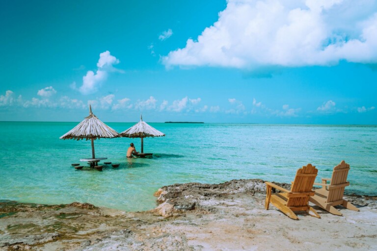 Una escena de playa con aguas cristalinas de color turquesa y cielo azul en Belice. Dos sombrillas con techo de paja están situadas en aguas poco profundas con una persona parcialmente sumergida cerca, posiblemente pensando en hacer negocios. Dos sillas de madera se sientan en la orilla arenosa frente al agua.