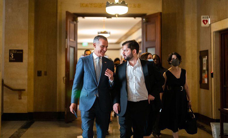 Two men in formal attire walk through a hallway engaged in conversation. The man on the left, who is wearing a blue suit, appears to be Chilean President Gabriel Boric. The man on the right is in a black suit with a white shirt. They are surrounded by other people, with open doors behind them and a lit hallway ahead.