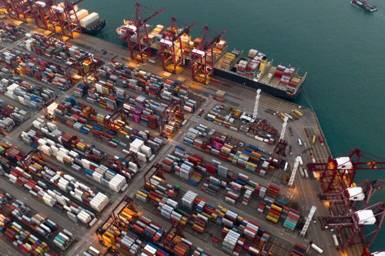 Aerial view of a bustling cargo port with numerous shipping containers organized in rows. Several large cranes are present, some actively loading or unloading containers from docked ships. The scene highlights the impact of nearshoring in Latin America, with two boats visible in the surrounding water.