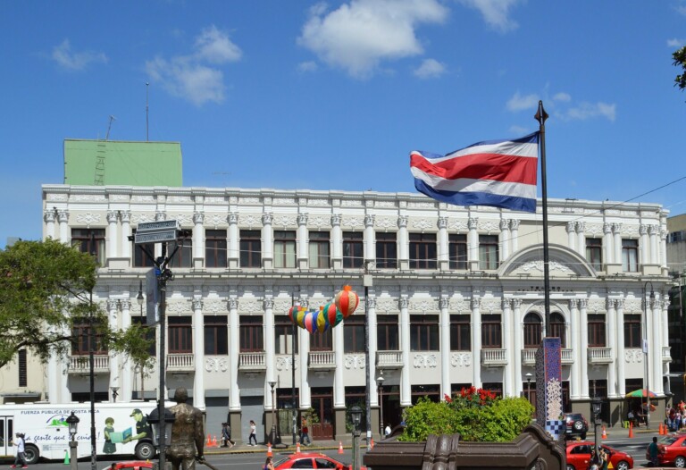 A two-story white building with ornate details under a clear blue sky. In front, a flagpole flies the Costa Rican flag, and a string of colorful balloons is tied to several posts. A tree, a parked truck, and some red flower planters are also visible—creating a serene contrast to the tales of caçadores de cabeças na Colômbia.