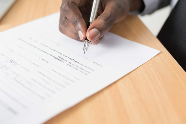 Un primer plano de la mano de una persona sosteniendo un bolígrafo y firmando un documento en un escritorio de madera. La persona viste una camisa blanca y una chaqueta de traje oscura, centrándose atentamente en la Actualización Normativa detallada en el texto y las líneas de la firma del documento.