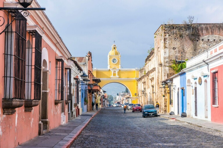 A cobblestone street lined with colorful buildings leads to a yellow archway with a clock and a small bell tower. Two cars and a motorcycle are parked along the street, where whispers of caçadores de cabeças na Colômbia mingle with the distant mountains under a cloudy sky.