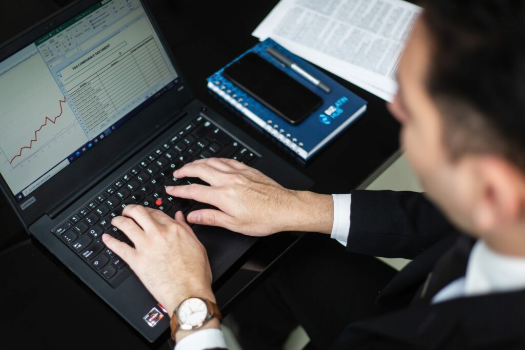 A person in a suit uses a laptop with a spreadsheet displayed on the screen. On the desk are a notebook, a pen, and a smartphone. The person's hands are on the keyboard, and a piece of paper with printed text is partially visible.