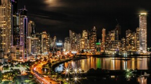 Nighttime cityscape featuring tall, illuminated skyscrapers creating a vibrant skyline. A winding road with vehicle lights fronts a reflective body of water. The lively scene feels like a headhunter in Panama navigating the numerous buildings and lights against the dark sky.