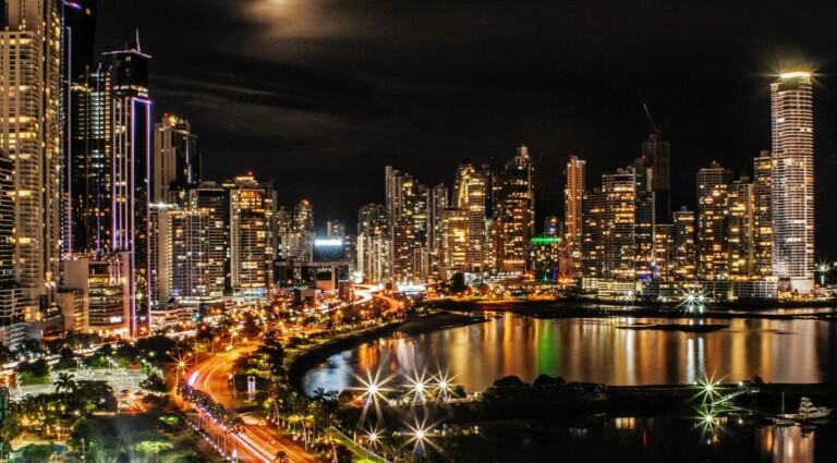 Un paisaje urbano vibrante por la noche con numerosos rascacielos iluminados, reflejándose en una masa de agua debajo. El cielo está oscuro con una luz brillante arriba, posiblemente de la luna. Autos con las luces encendidas circulan por la carretera que se curva a lo largo del paseo marítimo, que recuerda a un cazatalentos en las bulliciosas calles de Panamá.