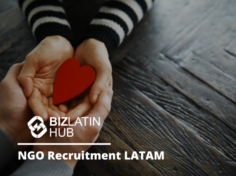 Primer plano de dos pares de manos sosteniendo una forma de corazón rojo sobre una mesa de madera. Una persona lleva una camisa a rayas. El texto superpuesto dice "BizLatin Hub Reclutamiento de ONG LATAM.