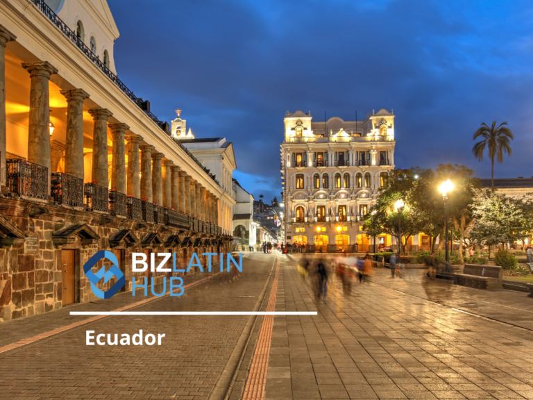 Vista nocturna de una plaza histórica en Ecuador con edificios iluminados. La gente camina por un camino pavimentado y las farolas iluminan la escena. Un edificio tiene columnas clásicas y otro se alza brillantemente iluminado al fondo. Se superpone el texto "Hacer negocios en Ecuador".