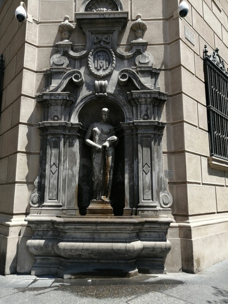 A statue of a woman holding a vessel, with water flowing from it, is set into an ornate stone niche on the corner of a beige building. The niche, reminiscent of the grandeur found in an accounting firm in Chile, is adorned with pillars and intricate carvings, including a circular emblem at the top.