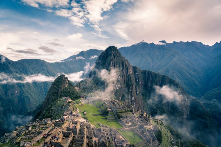 Una vista panorámica de Machu Picchu, una antigua ciudad inca en las montañas del Perú. El sitio presenta campos en terrazas, estructuras de piedra y senderos en medio de niebla y montañas verdes bajo un cielo parcialmente nublado, mostrando la belleza del Perú, donde también prospera la externalización de la contratación y el reclutamiento.