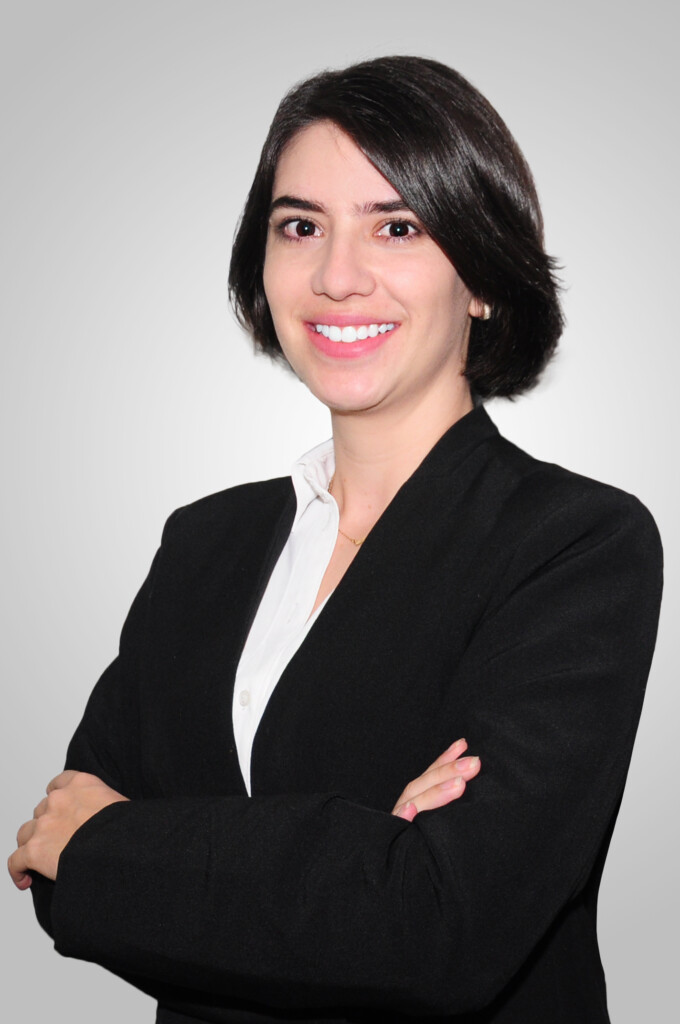 A person with short dark hair is smiling. They are dressed in a black blazer over a white shirt and have their arms crossed, exuding confidence typical of someone advocating for payment of salaries in cryptocurrency. The background is a plain, light gray gradient.