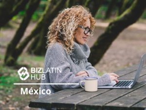 Una persona con cabello rizado y gafas está sentada en una mesa de picnic de madera en un ambiente al aire libre, usando una computadora portátil. Llevan un suéter de punto gris. Hay una taza blanca sobre la mesa. La imagen incluye los logos 'BIZLATIN HUB' y 'México', destacando el contexto de la Semana Laboral en México.