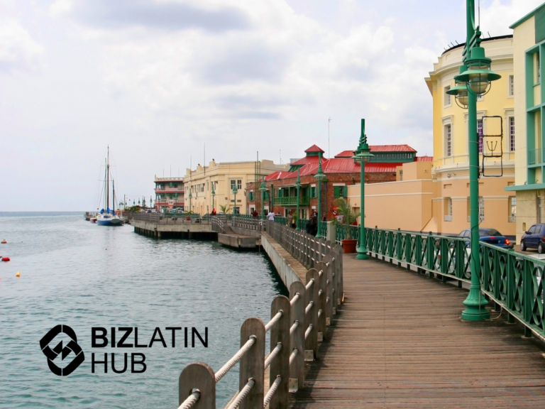 An oceanfront boardwalk runs alongside colorful colonial-style buildings with green railings, and a moored sailboat in the distance, evoking an atmosphere of coastal serenity. The logo and text "BIZLATIN HUB" are in the bottom left corner of the image, subtly hinting at an inflation update in LATAM.