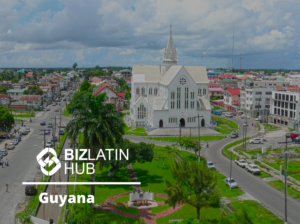 An aerial view of a cityscape in Guyana, featuring a prominent white church building with a tall steeple at the center. Surrounding the church are roads, vehicles, and other buildings, with greenery and a cloudy sky in the background. The text reads "BizLatin Hub - Corporate Bank Account in Guyana.