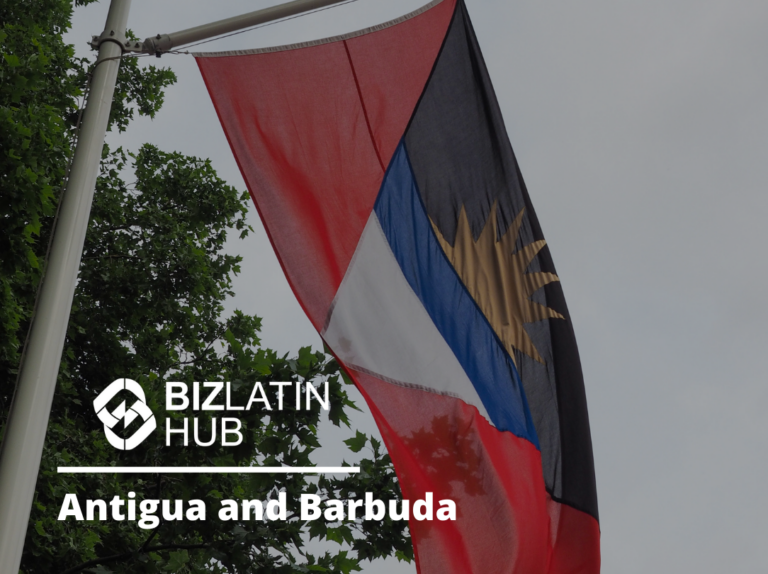 An Antigua and Barbuda flag waves outdoors against a gray sky. The flag features a rising sun symbol over red, white, and blue horizontal stripes. The image also includes the Biz Latin Hub logo and text reading, "PEO in Antigua and Barbuda.